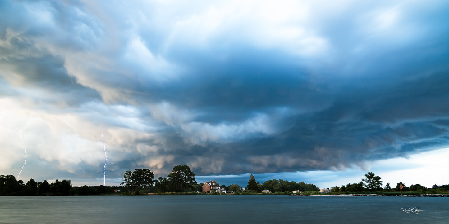 Electrified Horizon — Two lightning bolts striking behind a historic brick home on the shore of Piney Point Creek under a dramatic, heavy shelf cloud at twilight.