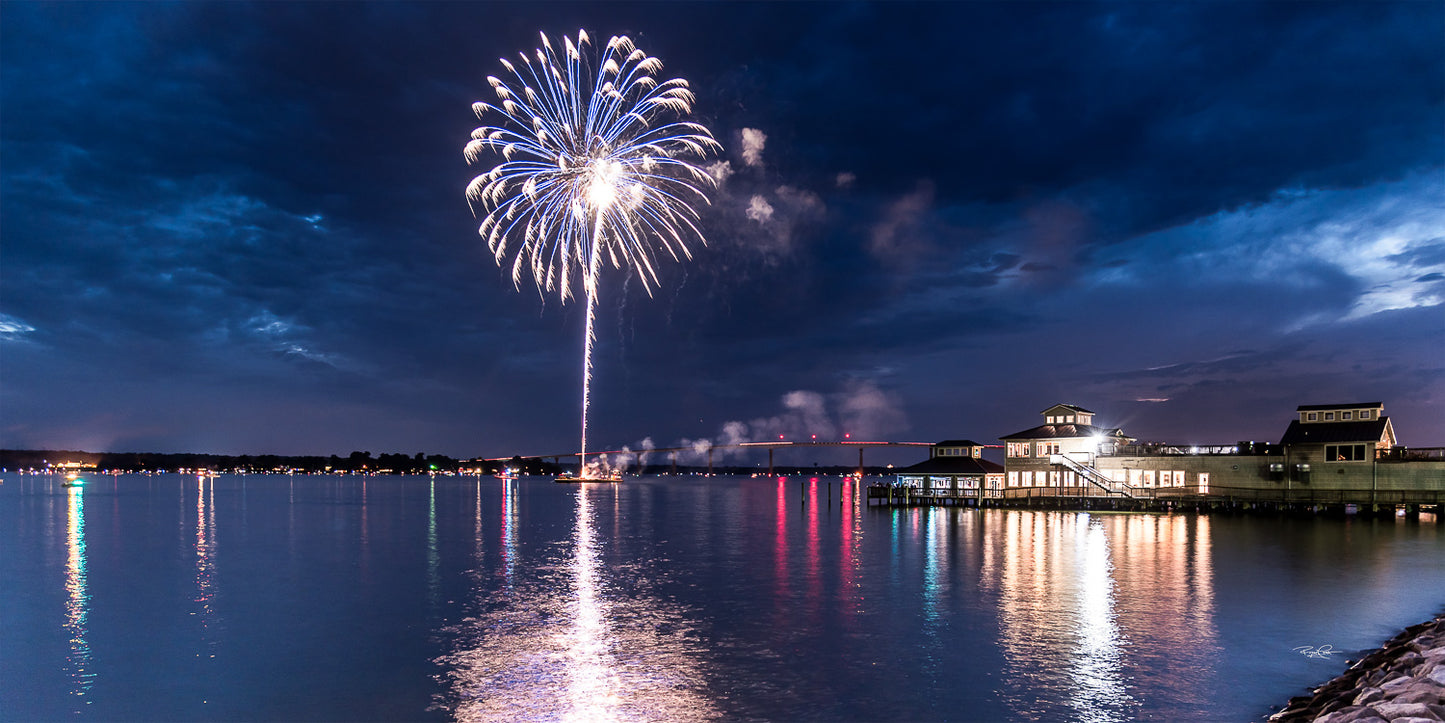 Solomons Spark — A large blue and white firework exploding over the water in Solomons Island, Maryland, with a waterfront restaurant and the Governor Thomas Johnson Memorial Bridge visible in the background.
