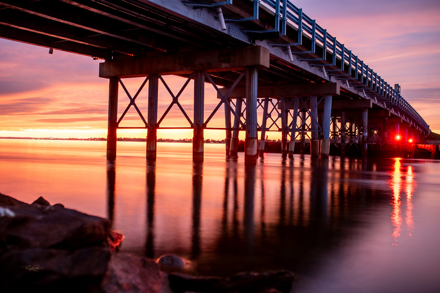 The Red Threshold — A long-exposure sunset photograph taken from beneath the St. George's Island bridge, featuring deep orange and pink sky colors reflected in glassy water with the bridge's red navigation lights glowing on the right.