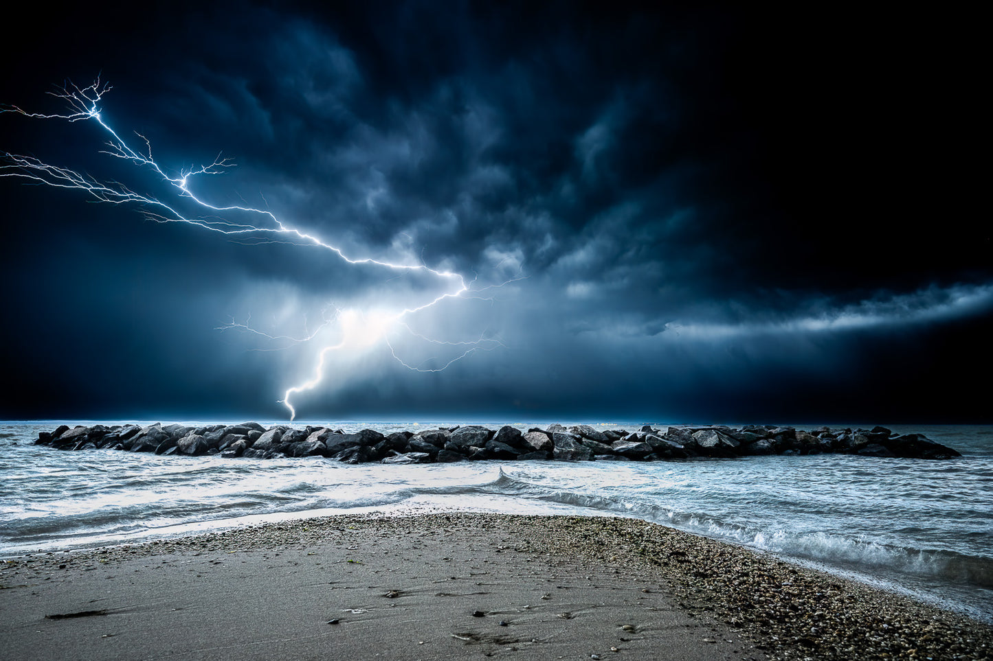 The Electric River — A dramatic long-exposure photograph of a bright lightning bolt striking the horizon behind a rock jetty on the Potomac River, under a dark, swirling storm sky at night.