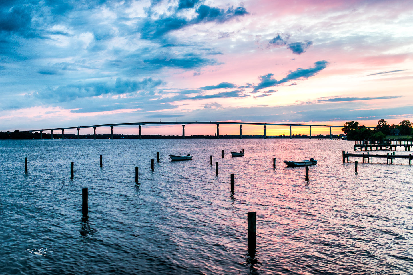 Evening Passage — A wide-angle view of the Governor Thomas Johnson Memorial Bridge at sunset, with a vibrant pink and blue sky reflected in the Patuxent River and small boats anchored in the foreground.
