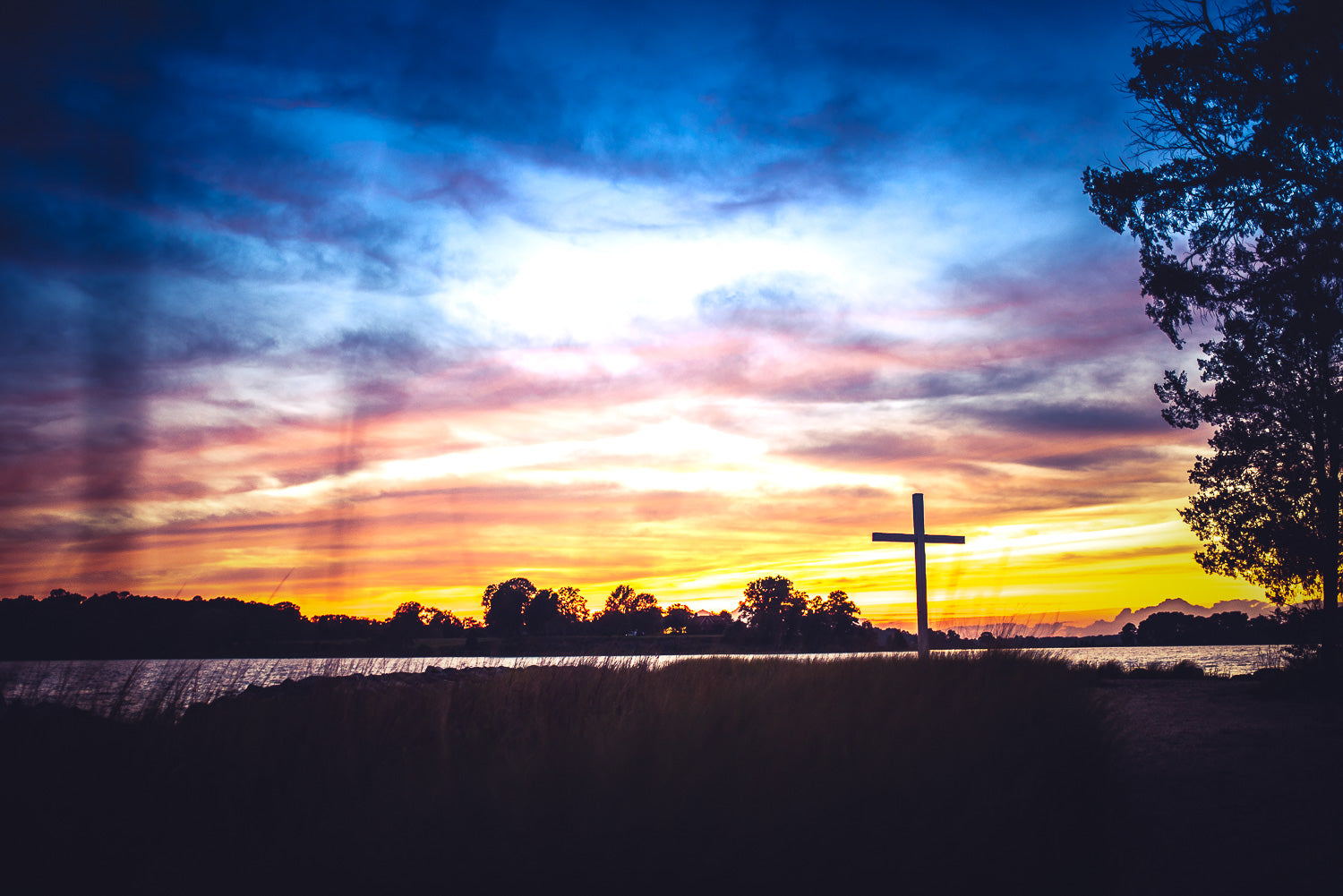 Guardian of the Point — A silhouette of a wooden cross on the shore of Priest's Point at St. Mary's College, set against a vibrant purple, orange, and blue sunset reflecting over the St. Mary's River.