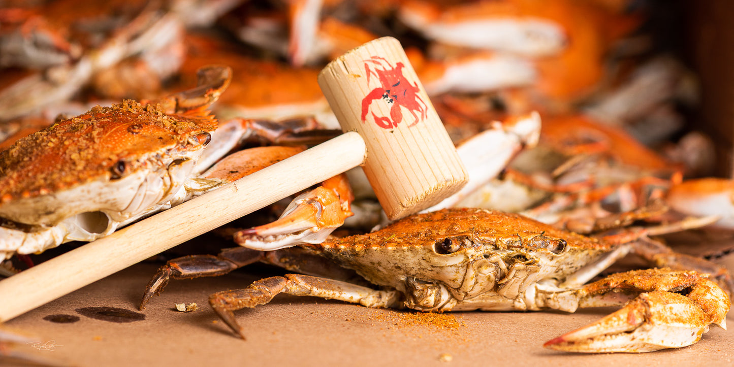 The Seasoned Strike — A close-up of steamed Maryland blue crabs heavily seasoned with red spice, with a wooden crab mallet resting across the foreground.