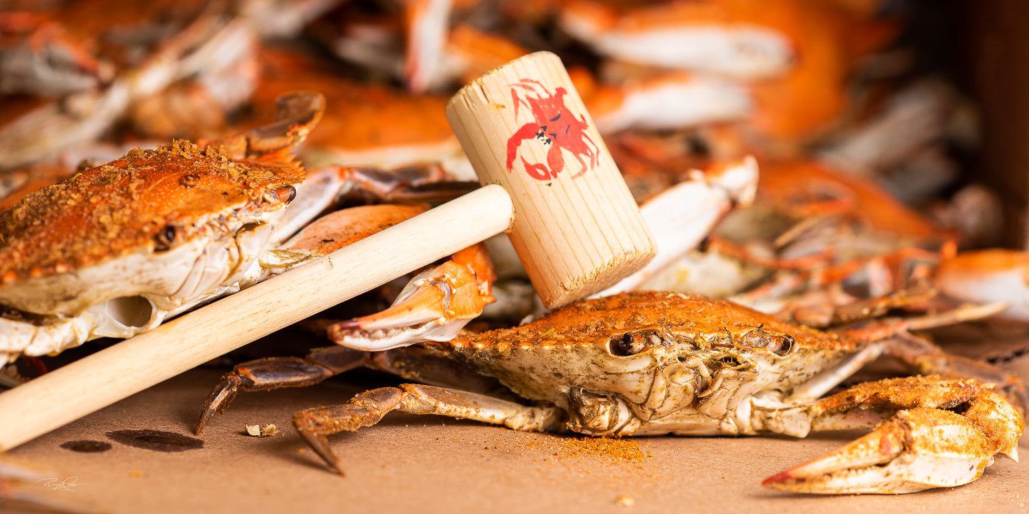 The Seasoned Strike — A close-up of steamed Maryland blue crabs heavily seasoned with red spice, with a wooden crab mallet resting across the foreground.