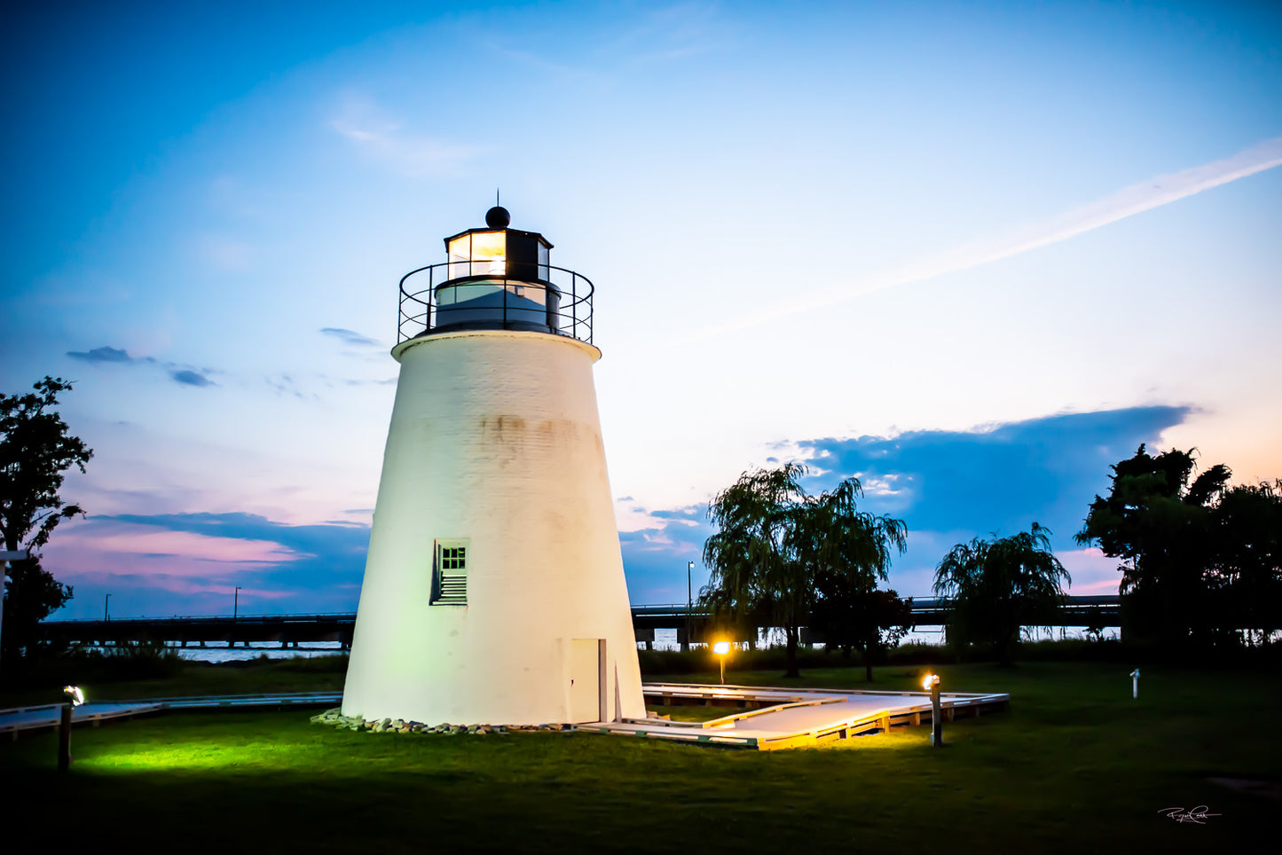 The Evening Sentinel — A white conical lighthouse at Piney Point, Maryland, illuminated at twilight with a soft pink and blue sky in the background and glowing walkway lights in the foreground.