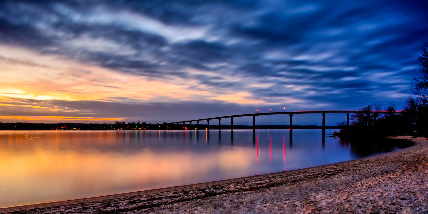 The Still Crossing — A panoramic long-exposure twilight photograph of the Governor Thomas Johnson Memorial Bridge with glass-smooth water, warm sunset glow transitioning to cool blue, and red bridge light reflections.
